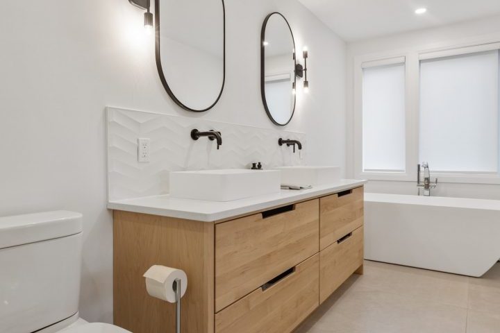 A bright and spacious bathroom with a double vanity featuring two sinks and chrome faucets. There are mirrors with wall sconces above each sink and a bathtub next to the vanity. A toilet is located on the opposite side of the room. The bathroom has white walls and beige floor tiles.