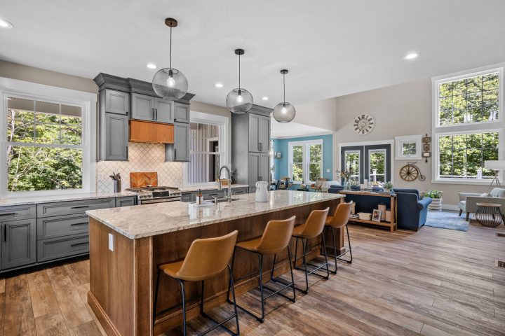 A modern kitchen showcasing a center island with a breakfast bar overhang. The island countertop is light marble with a waterfall edge for a polished look. Four brown leather bar stools with backs provide additional seating beneath the overhang. Gray cabinets for ample storage, and hanging pendant lights for additional lighting solutions.